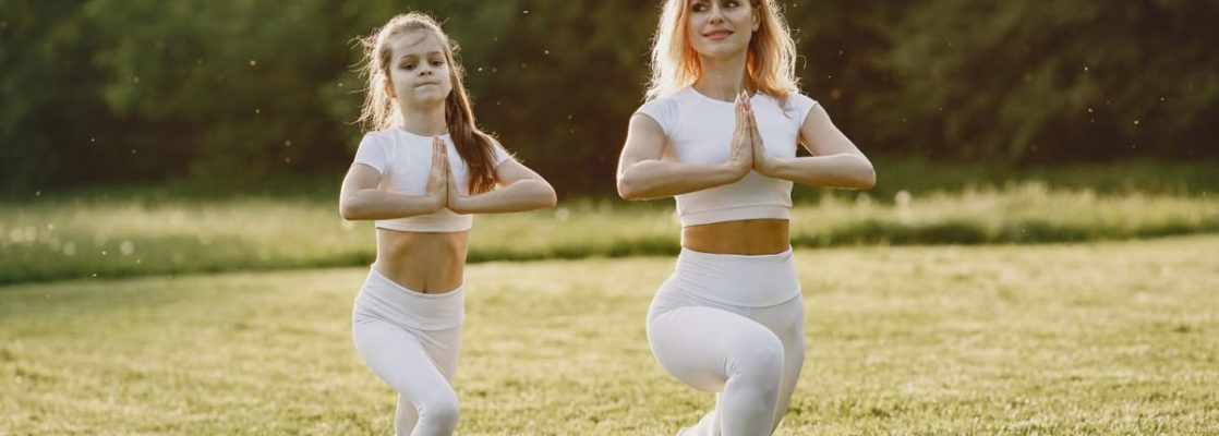 A mother and daughter practicing yoga together in matching outfits in a sunny park.